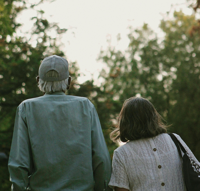 Photo of an elderly couple from behind while they stand outside looking into the forest